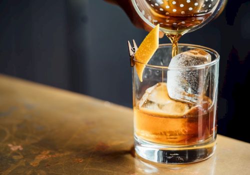 A glass of whiskey on the rocks being garnished with an orange twist and a large ice cube, with a carafe pouring above it.