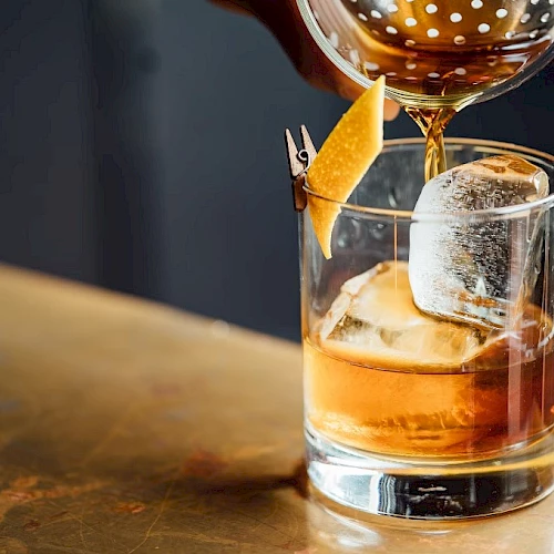 A glass of whiskey on the rocks being garnished with an orange twist and a large ice cube, with a carafe pouring above it.