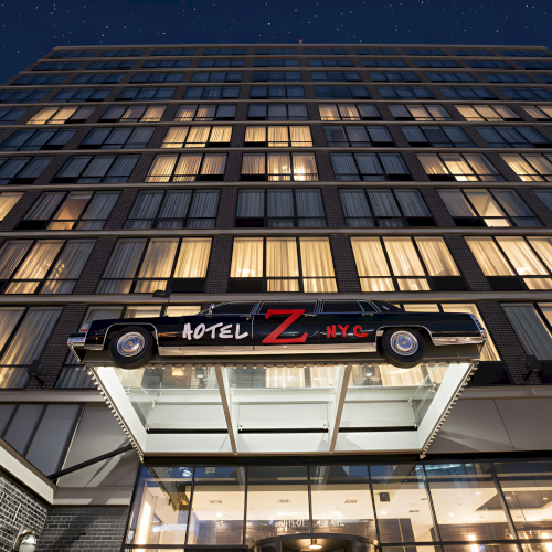 A multi-story hotel building with a car featuring "Hotel Z NYC" on its side above the entrance, set against a night sky.