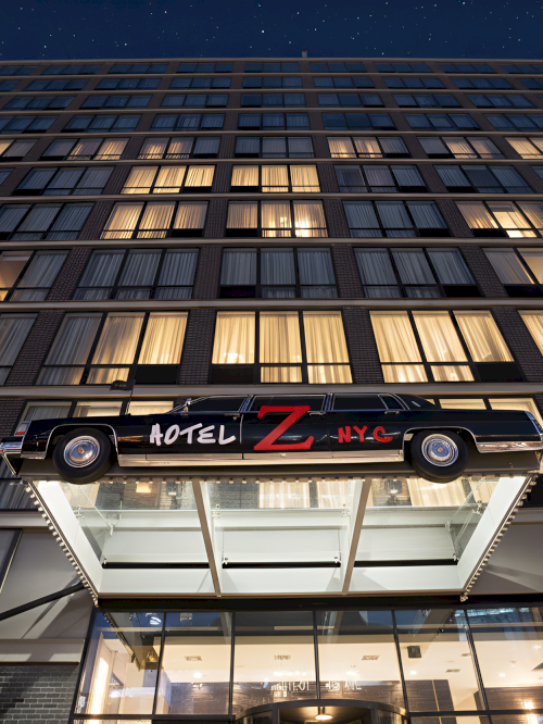A multi-story hotel building with a car featuring "Hotel Z NYC" on its side above the entrance, set against a night sky.