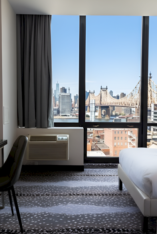 A modern hotel room with two beds, a desk, a chair, and a view of a bridge through large windows, creating a stylish atmosphere.