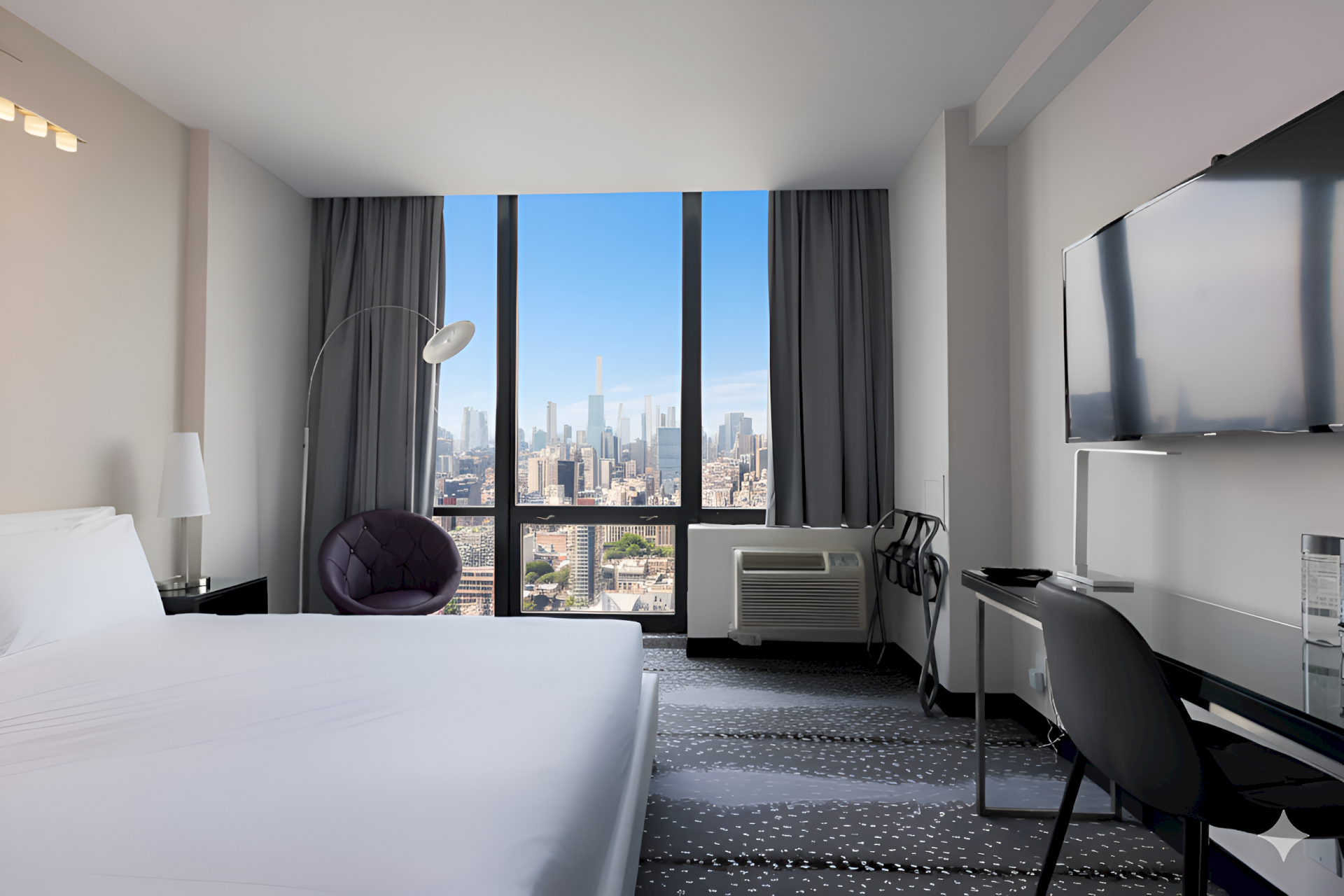 A modern hotel room with a city skyline view through tall windows, dark curtains, a bed, and a desk with a large monitor.