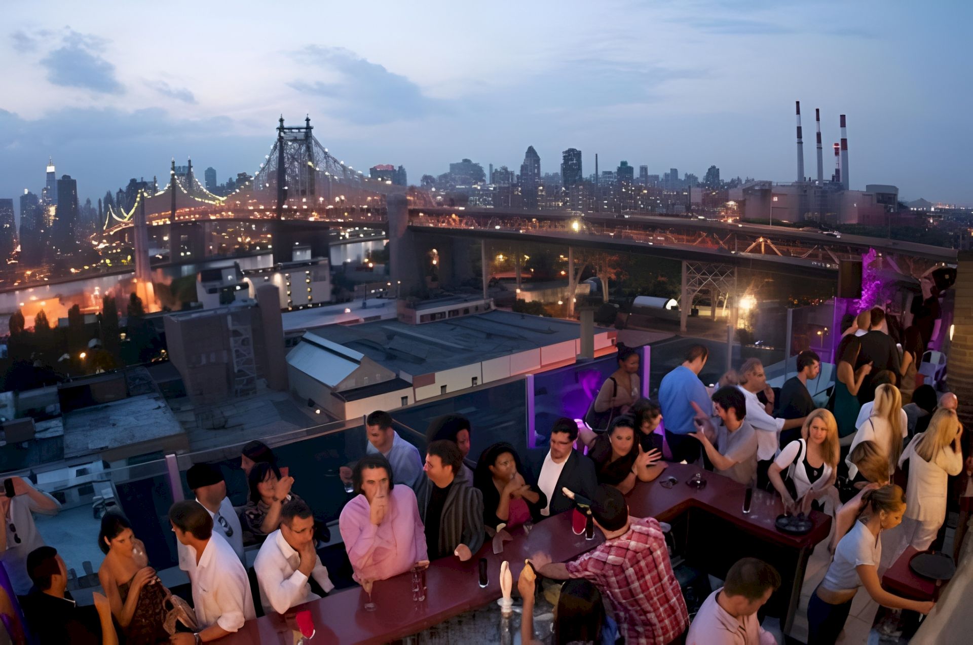 People mingle on a rooftop terrace at dusk overlooking the Brooklyn Bridge and a glowing city skyline.