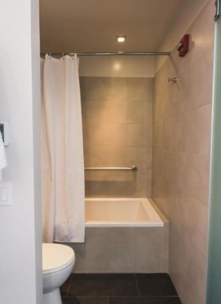 A small bathroom with a bathtub and shower curtain, a toilet, light gray tiled walls, and a robe or bathrobe hanging nearby, all inside a compact space.