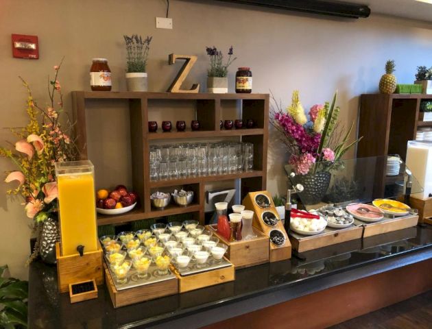 A breakfast buffet setup with juice, yogurt cups, pastries, and fruit on a dark wooden counter, decorative flowers, and shelves in back.