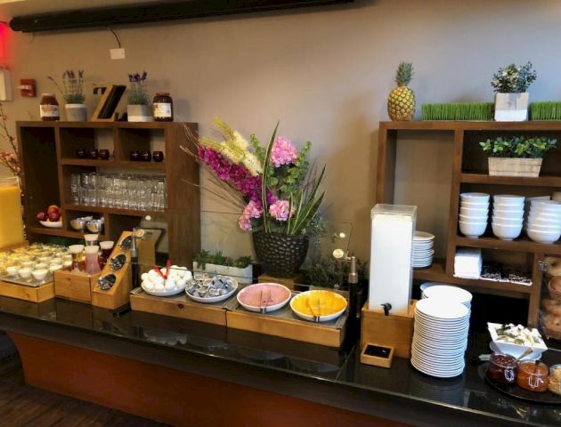 A cozy breakfast buffet with breads, pastries, fruit, yogurt, cereals, and hot plates from a wooden self‑serve counter, plus shelves of bowls and decor.