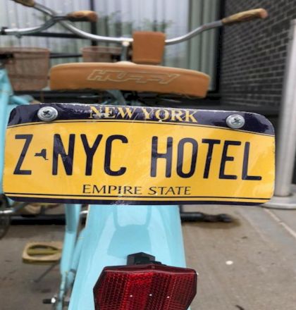 Vintage New York plate on mint blue bike, reads “Z NYC HOTEL” with Empire State text, orange seat, and red tail light in an alley.