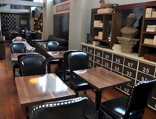 A cozy cafe with dark wooden tables and black-studded chairs, shelves and busts along the wall, and a chalky number-printed backdrop.