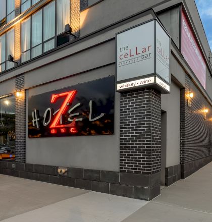 A modern street-side bar/restaurant with red seating along the wall, large illuminated sign, brick accents, and a sign reading “the CELLAR” on the corner. Top it at 140 characters, ending with a period.