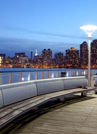 A nighttime city skyline along a waterfront boardwalk with curved benches and modern street lamps, reflecting in calm water near the skyline.
