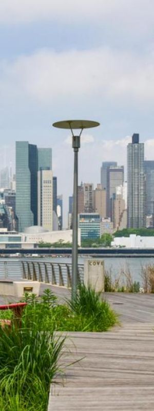 Two red Adirondack chairs face a modern city skyline across a river, on a wooden boardwalk with greenery and a lamp post, under a partly cloudy sky.