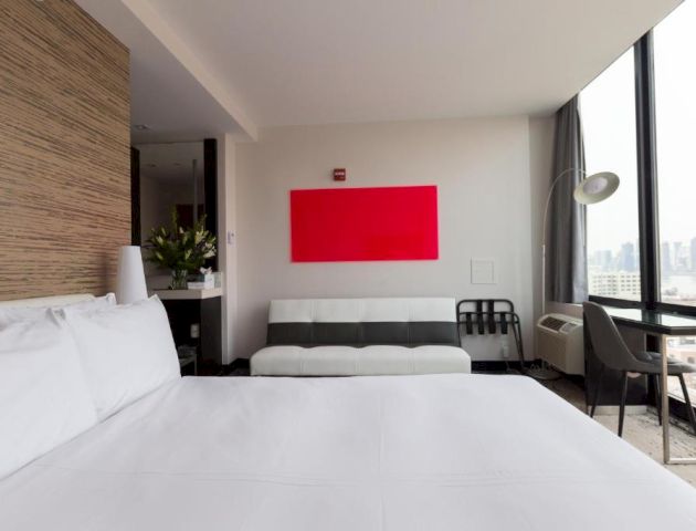 A modern hotel room with a large bed, a gray sofa, a red abstract wall art, a desk by the window, and city views outside.