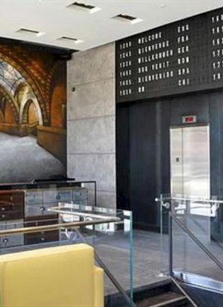 An airy, modern train station lobby with glass railings, a yellow bench, a large mural of a curved arched tunnel, and an ascent/exit area with digital boards.