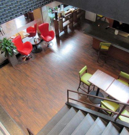 A modern lounge and cafe area with wooden floors, red accent chairs, a long counter, and several green chairs around tables.