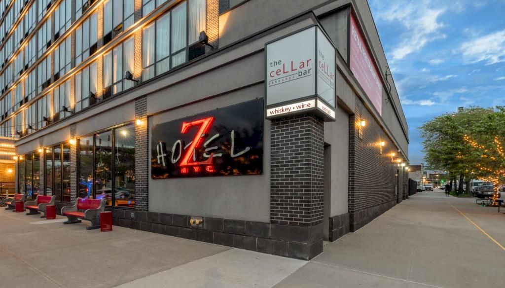 A modern street view of The Cellar Bar at a corner, with red seating along the sidewalk, neon signage, brick and glass facade, and a clear blue sky.