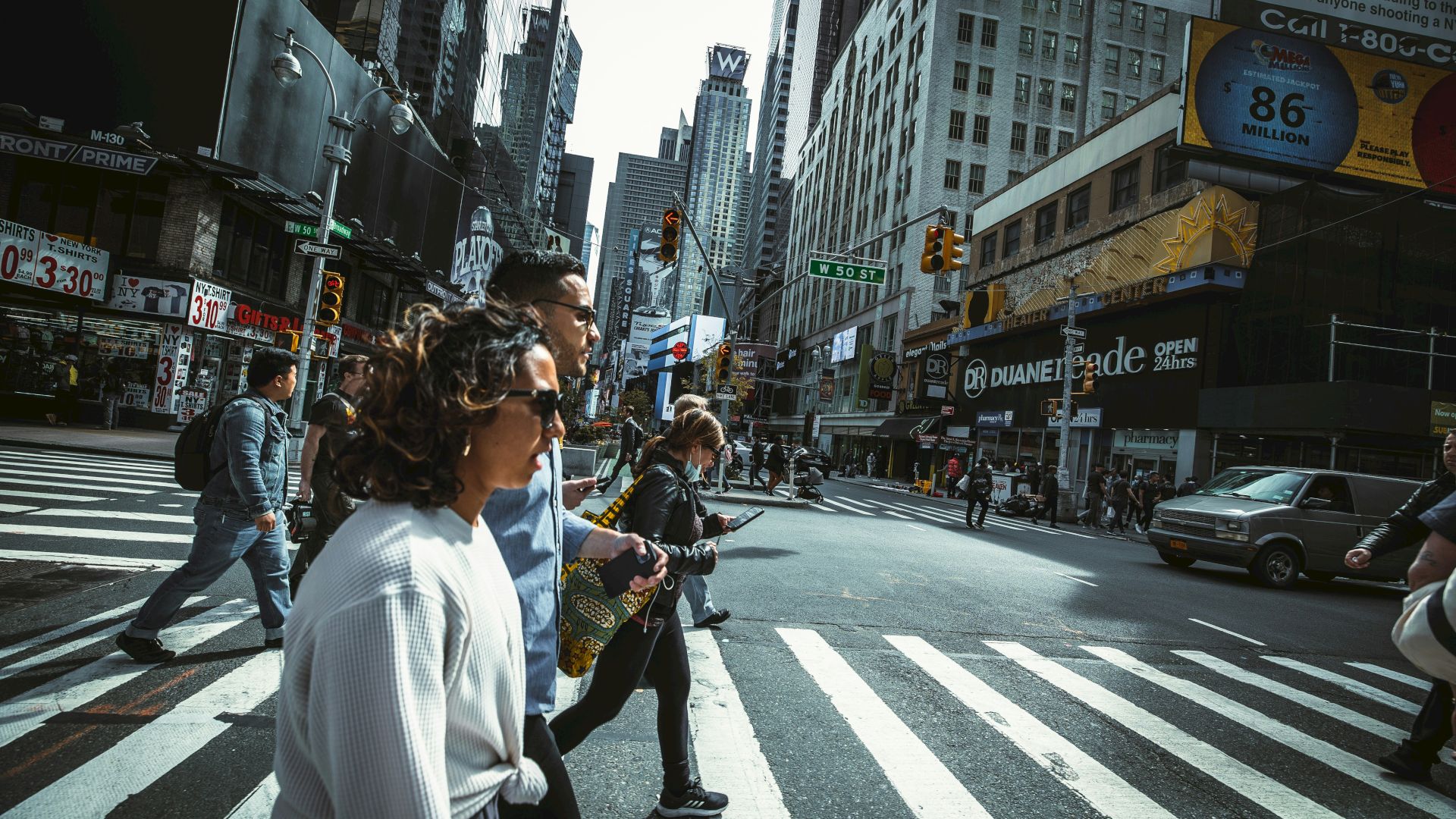 People cross a busy city street at a crosswalk; tall buildings line the avenue, storefronts, and cars in the background.