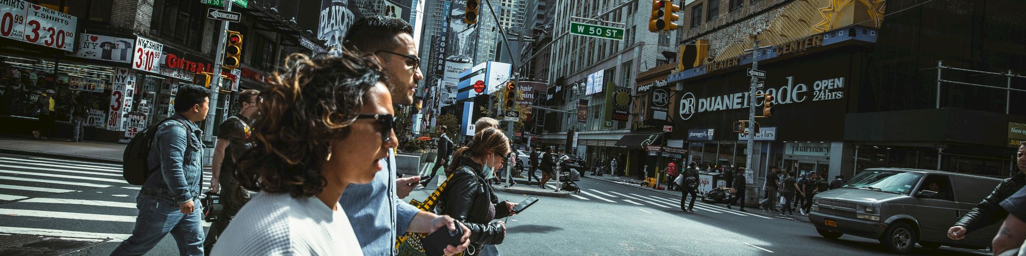 People cross a busy city street at a crosswalk; tall buildings line the avenue, storefronts, and cars in the background.