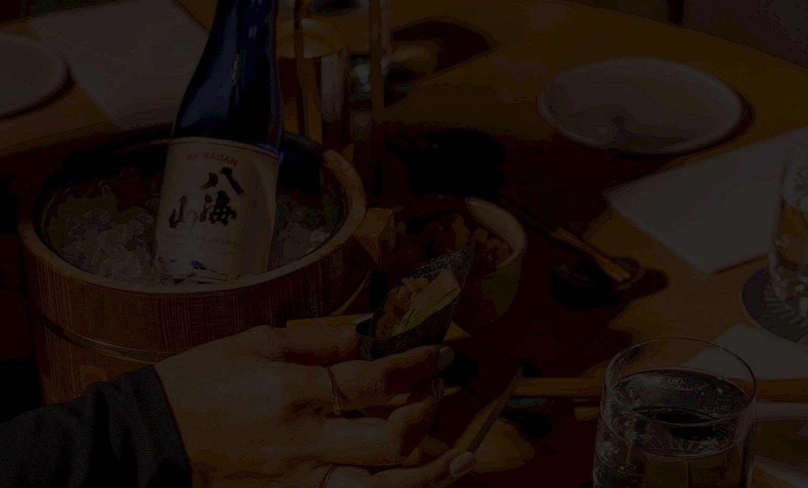 A person toasts with a bottle of sake in a wooden bucket, surrounded by glasses and dishes at a dim table, celebrating together.