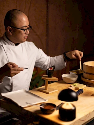 A person, likely a tea ceremony host, sits at a wooden table with bowls, a teapot, and utensils, carefully preparing tea, focused and serene.