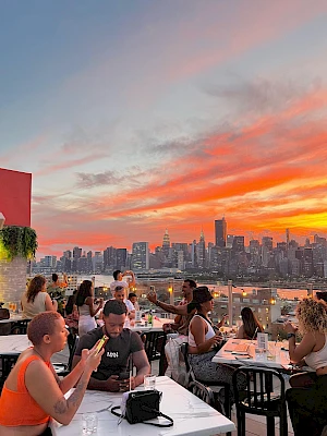 A rooftop dinner scene with friends at white-clothed tables, overlooking a bright sunset and a city skyline in the background, animated chatter.