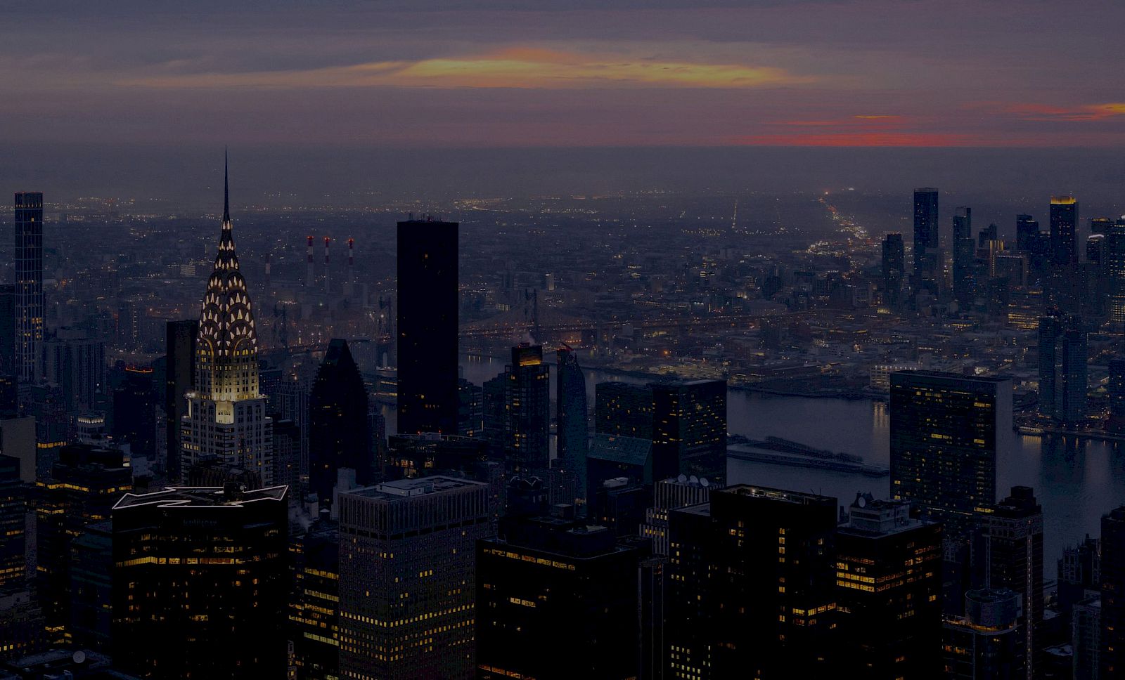 A nighttime city skyline with tall skyscrapers and a distant river, softly lit as the sunset fades behind the horizon.