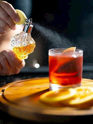 A hand sprinkles powdered sugar or salt onto a glass perfume bottle beside a red cocktail with a lemon twist on a wooden tray.