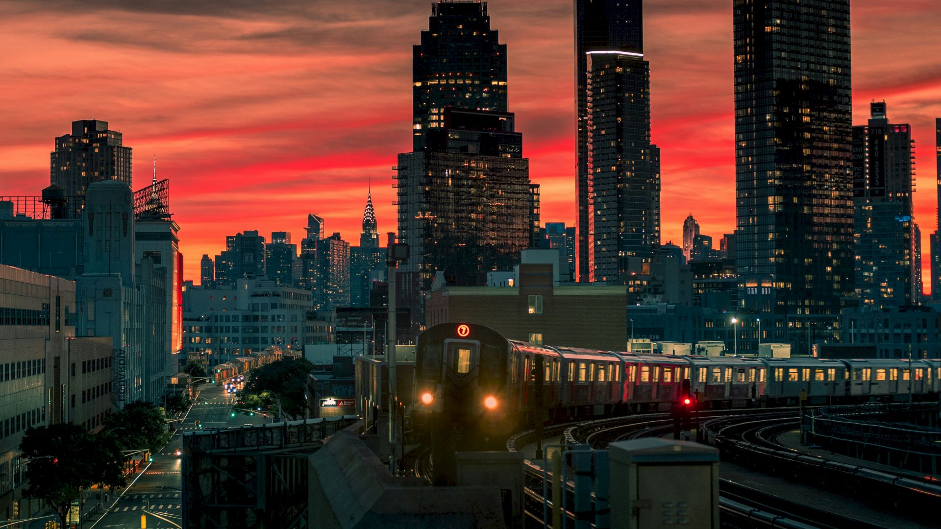 A sunlit city skyline glows crimson as a train glides along elevated tracks, weaving through tall buildings against a fiery sunset backdrop.