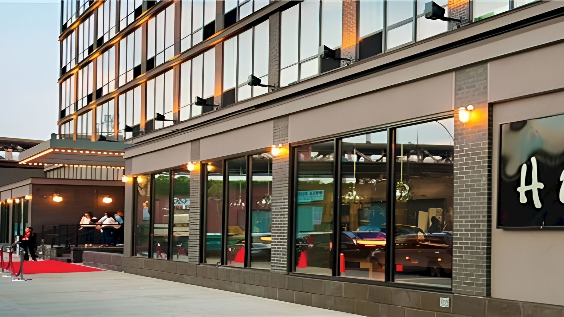 A modern glass-front building with people at an entrance, warm lights, and a red carpet leading into a stylish venue or hotel lobby.