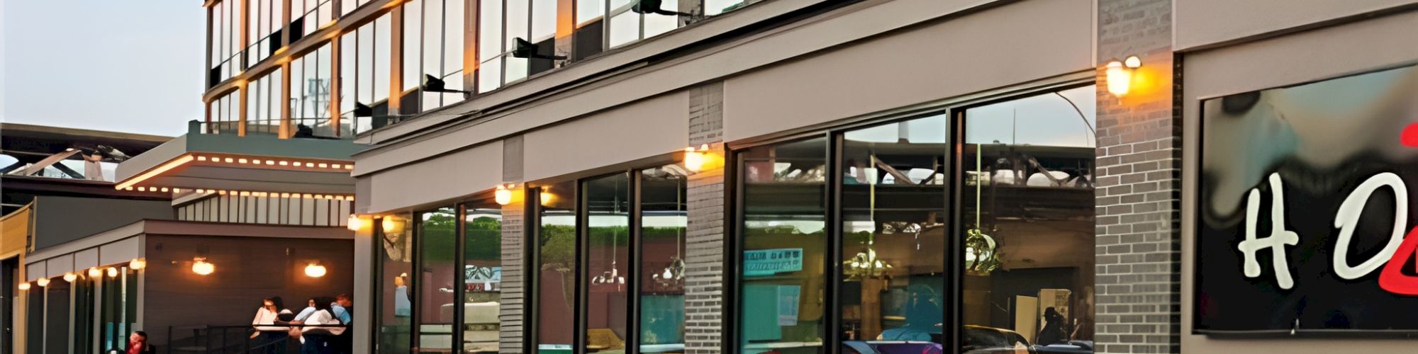 A modern glass-front building with people at an entrance, warm lights, and a red carpet leading into a stylish venue or hotel lobby.