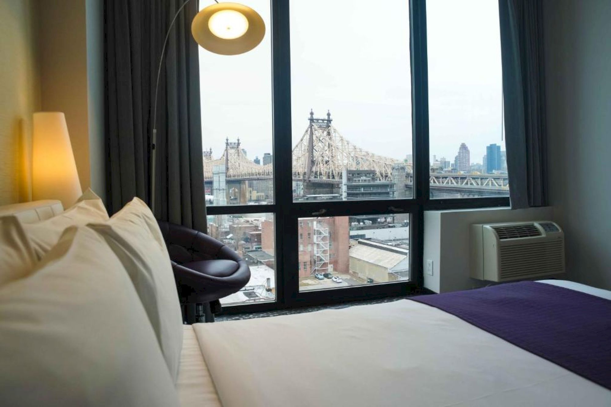 A cozy hotel room with a bed, nightstand lamp, a chair, and a large window overlooking a city with a bridge in the distance.
