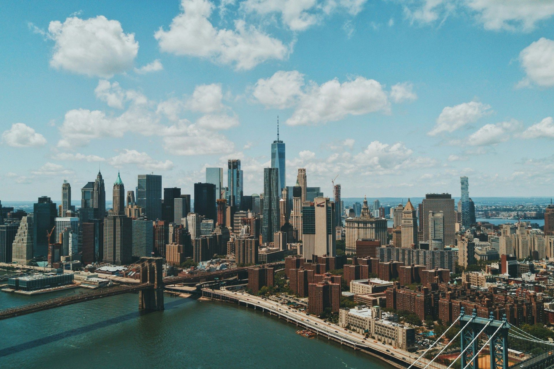 A panoramic New York City skyline with skyscrapers, a riverfront, and a bright, partly cloudy sky, showcasing tall modern buildings.