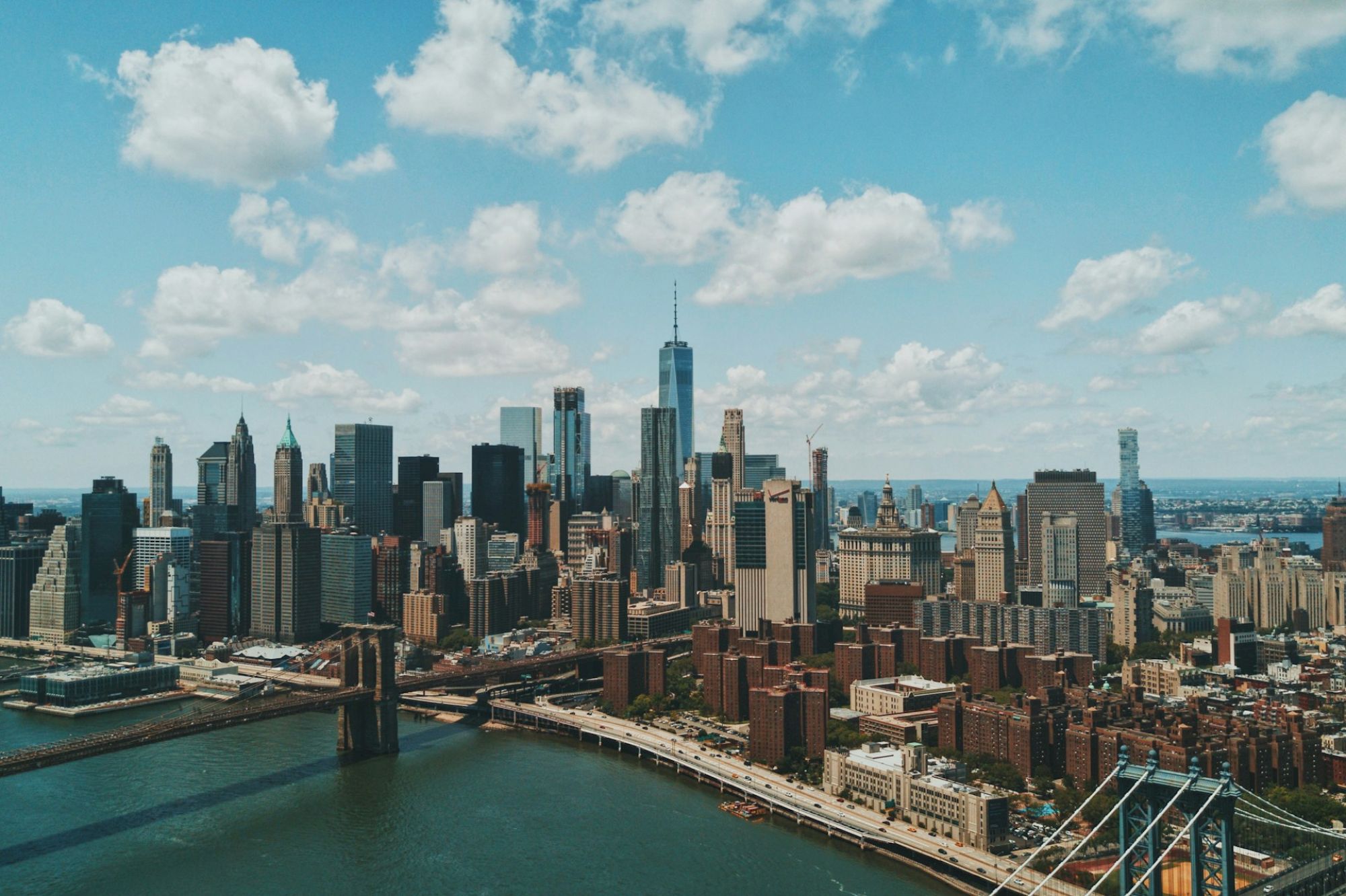 A panoramic New York City skyline with skyscrapers, a riverfront, and a bright, partly cloudy sky, showcasing tall modern buildings.
