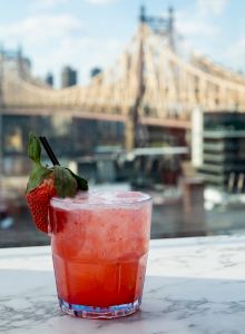 A pink-red cocktail with a strawberry garnish on a marble surface, city skyline and a bridge in the background, hinting at an urban rooftop view.
