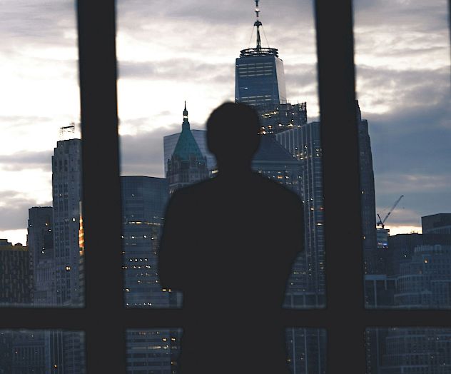 A person stands by a large window looking out over a city skyline at dusk, silhouetted against tall buildings. End.