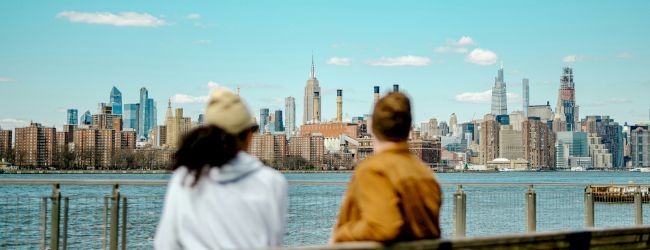Two people sit on a bench by the water, facing a city skyline across the harbor on a clear day.