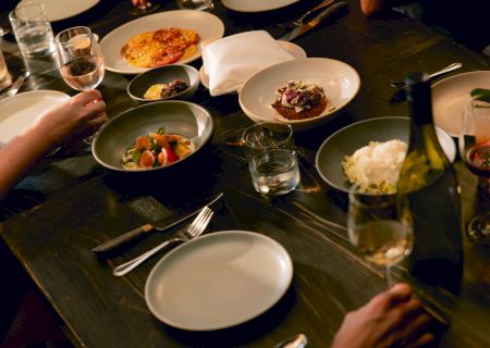 A dinner table with several plates of food, a bottle of champagne, wine glasses, and hands reaching for dishes. Noting a white plate and various small bowls of food.