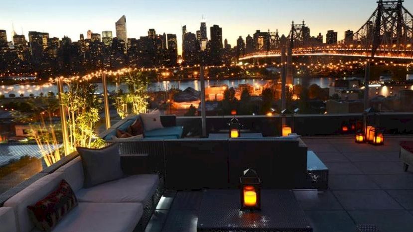 A city skyline at dusk from a stylish rooftop lounge with comfortable seating, warm lanterns, and a lit bridge in the background.